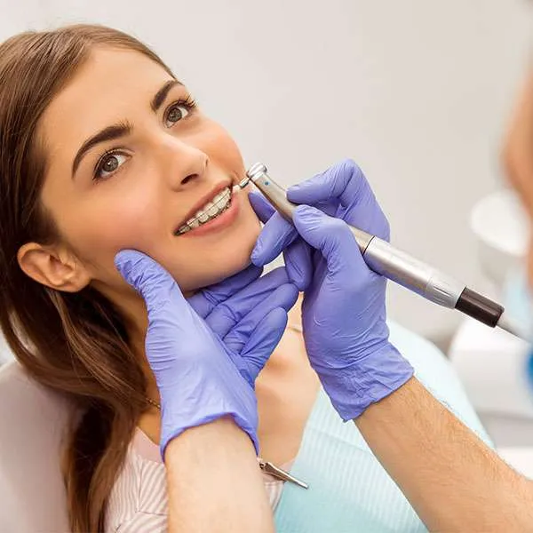 A young woman with braces is sitting in a dental chair while a dentist wearing purple gloves uses a dental tool to examine or clean her teeth.